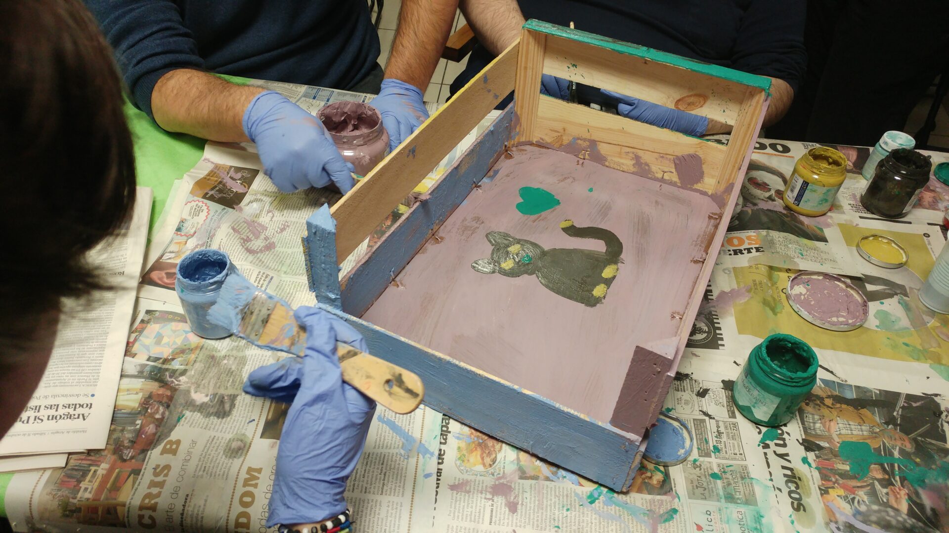 Taller de fabricación de juguetes para animales con material reciclado, aparece una caja de madera que están pintando para convertirla en la cama de un gato